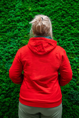 A woman with a ponytail in a red hoodie stands with her back to the camera against a background of decorative moss