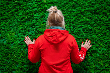 A woman with a ponytail in a red hoodie stands with her back to the camera against a background of decorative moss
