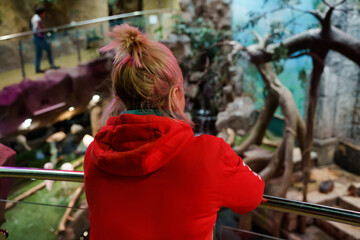 A woman in a red hoodie stands on the second floor and looks at the landscaping from below at the zoo