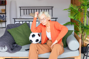 beautiful elderly senior woman in cosy apartment watching a football match, beautiful gray haired woman with soccer ball, concept of active old age