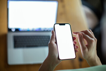 Woman holding smartphone with blank screen in focus laptop in background