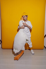 photo session of a boy in a yellow hat and white T-shirt with a toy bird goose hugging standing on a yellow paper background in a studio
