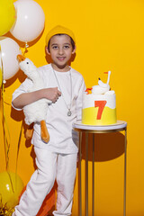 photo session of a boy in a yellow hat and white T-shirt with a toy bird goose hugging standing on a yellow paper background in a studio. standing next to a cake with a goose on it