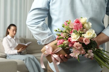 Man hiding bouquet of flowers and present for his beloved woman indoors, closeup
