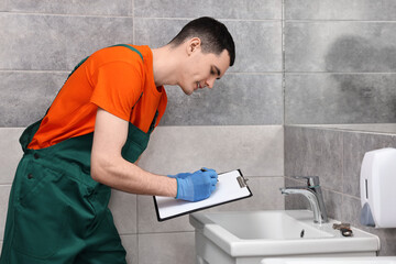 Young plumber writing results of examining sink in bathroom