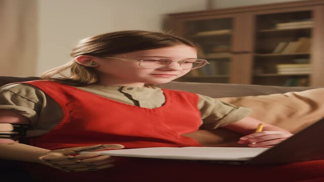Vertical side footage of diligent Caucasian homeschooler girl with bionic arm studying online and watching educational video or lesson on laptop