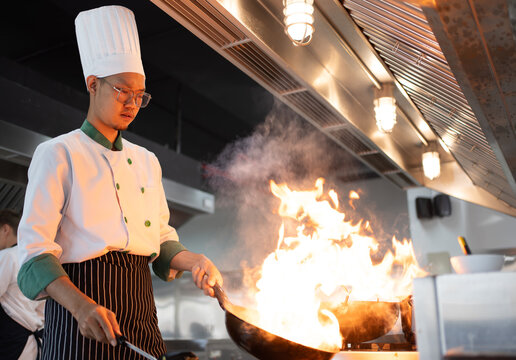 Restaurant Kitchen Action. Chef cook food with fire at kitchen restaurant Cook with wok at kitchen Chef male in uniform hold wok with fire. Сhef in the professional kitchen with a frying pan..