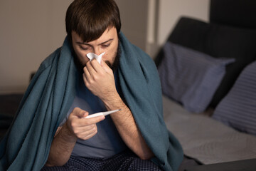 Sick young man sitting blanket sofa, taking his temperature thermometer, sneezing napkin home....