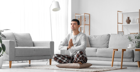 Young man in pajamas meditating on pillow at home - obrazy, fototapety, plakaty