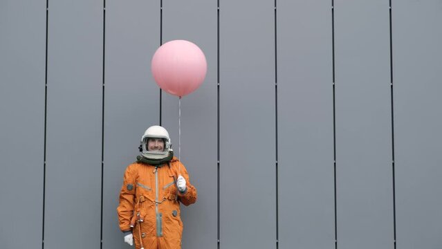 Happy astronaut wearing orange space suit and astronaut helmet holding pink helium balloon on gray background