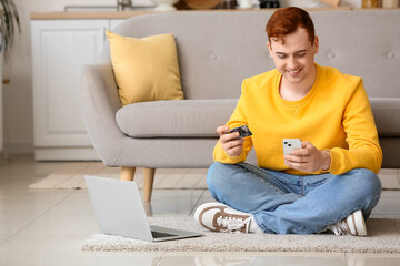 Handsome young man with credit card and mobile phone sitting on floor near sofa while shopping...