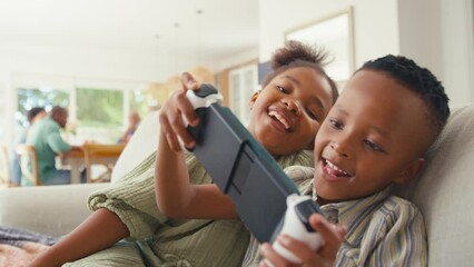 Boy and girl sitting on sofa playing with handheld gaming device at home with rest of family in background - shot in slow motion