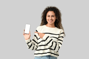 Fototapeta premium Young African-American woman pointing at mobile phone with blank screen on grey background