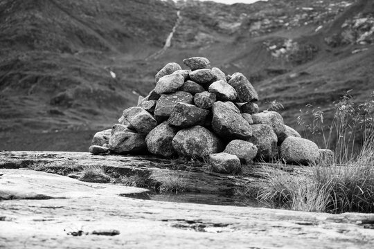 A carefully stacked cairn stands on a rocky terrain with lush greenery and a waterfall background. Black and white photography.