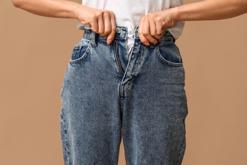 Beautiful young Asian woman buttoning up stylish jeans on brown background, closeup - obrazy, fototapety, plakaty