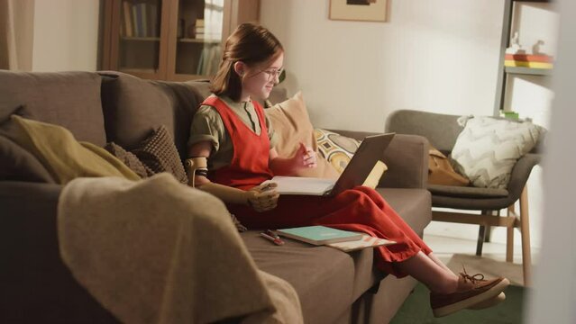 Side full footage of Caucasian little schoolgirl with prothetic arm answering during online lesson while sitting on sofa with notebooks in cozy living room