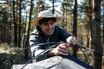 vertical portrait Smiling young man with hat assembling tent poles at campground during summer sunset