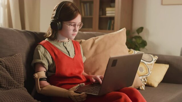 Side footage of Caucasian homeschooler girl with bionic arm wearing glasses and headphones learning online at home