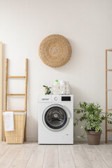 Modern washing machine with basket, houseplant and ladder near white wall. Interior of home laundry...