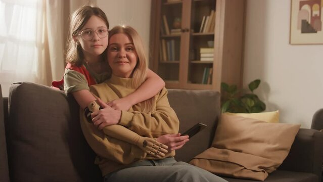 Medium portrait of Caucasian single-parent family of little daughter with prosthetic arm who hugging her smiling mother from behind and looking at camera at home