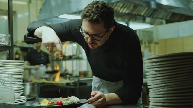 Master chef in uniform leaning on kitchen table and adding olive oil to fish served with vegetables on plate when working in fine dining restaurant