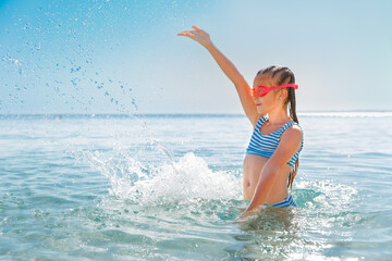  Childhood water fun: girl with swimming goggles splashing water in the sea.