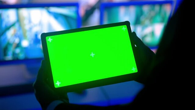 Man security guard officer in monitoring center holding tablet with chroma key green screen. Computers and big digital screen show cctv cameras footage.
