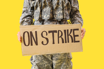Protesting male soldier holding placard with text ON STRIKE against yellow background, closeup