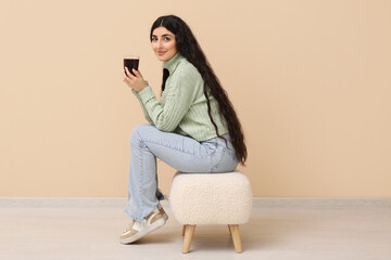 Beautiful young woman with glass cup of hot tea sitting on pouf near beige wall