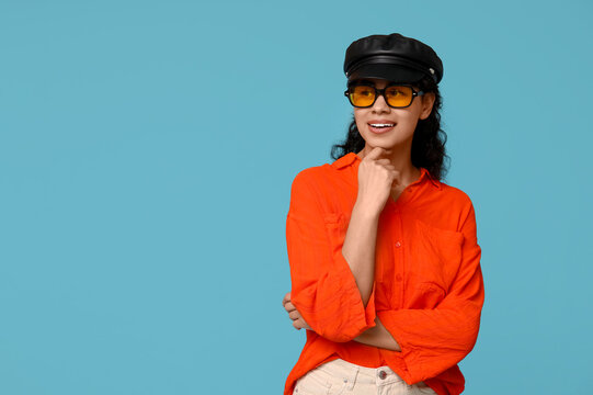 Stylish African-American woman wearing sunglasses and hat on blue background