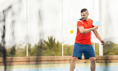 An adult man in sportswear is playing a game of pickleball on an outdoor court.The man is making a backhand with the racket to hit the ball.The photo is seen through the net of the track. - Powered by Adobe