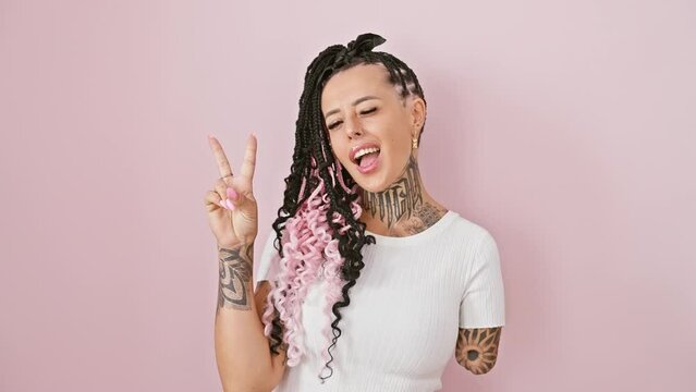 Exuberant hispanic amputee woman shows victory sign, winking and smiling over pink isolated background