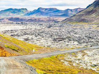 A road winds beside a vast, solidified lava field under a cloudy sky, with rugged hills in the background, depicting the stark terrain of Reykjanes Peninsula in Iceland.