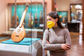 Female museum visitor in protective face mask examining with interest ancient stringed musical...