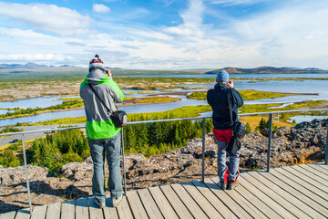 Two visitors stand on a wooden platform admiring the expansive view of Thingvellir National Park in Iceland, with clear skies and serene waters in the backdrop.