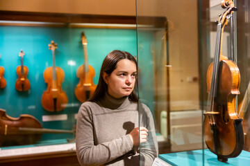 Female museum visitor examining with interest ancient stringed musical instruments displayed on exhibition ..