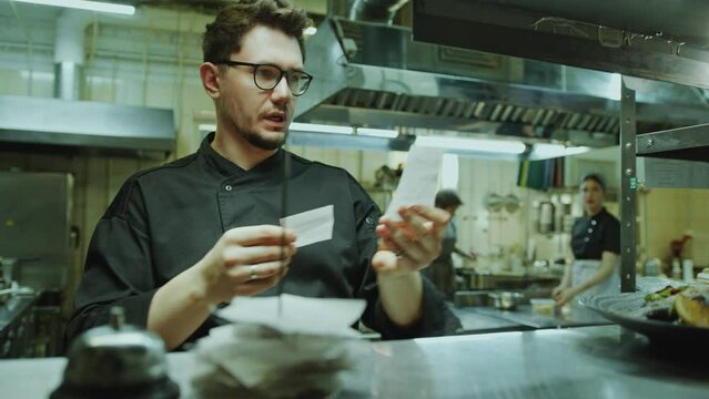 Chef taking order tickets from metal rack, reading them and telling what to cook to kitchen crew during workday in restaurant