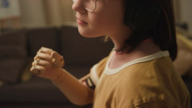 Tilt closeup of cute Caucasian teenage girl with prosthetic arm dancing while listening to music in headphones at home