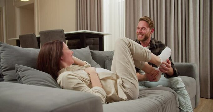 Happy pregnant brunette woman with a bob hairstyle in a white T-shirt plays with her blond husband in a plaid shirt while he tickles her on a gray sofa in a modern apartment at home
