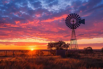 Schilderijen op glas Colorful Australian outback sunset landscape with a windmill, water tank and gumtrees and a firey sky with clouds with a sunburst. © Inge