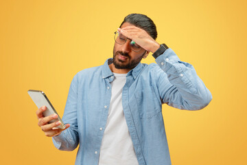 Confused man checking time on smartphone, yellow backdrop