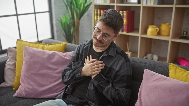 Heartwarming portrait of a young hispanic man, gleaming in his glasses, gratefully beaming while sitting on a cozy sofa at home. hands to chest, an image of good health and high spirits.