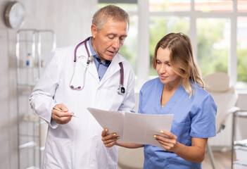 Two fellow doctors, adult woman and senior man, standing in medical office with papers in hands, focused on discussing of clinical diagnosis of patient - obrazy, fototapety, plakaty