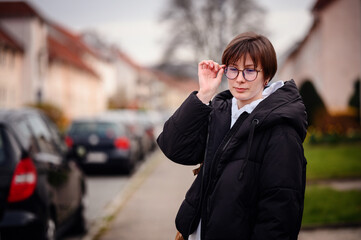 A contemplative young woman is adjusting her glasses, standing on a quiet city street, with houses...