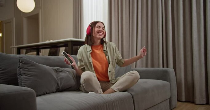 A happy brunette girl with a bob hairstyle and wireless red headphones sits on a gray sofa and listens to cheerful rhythmic music using her phone in a modern apartment having fun on vacation