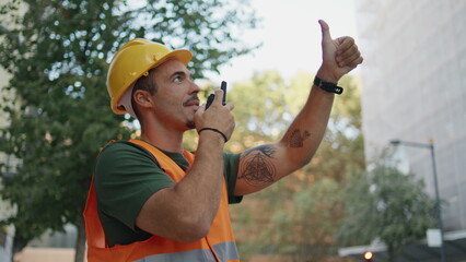 Uniform engineer showing thumb talking walkie-talkie at urban district closeup