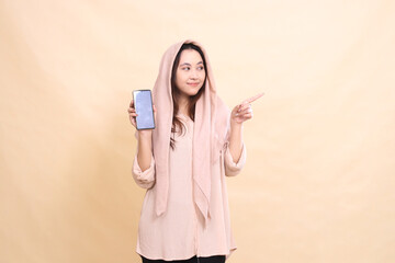 indonesia woman smiles broadly to the left with her left pointing hand and holds the cellphone gadget screen. young beautiful for business, technology, fashion