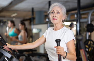 Slender athletic mature woman during cardio exercises on elliptical simulator in fitness center....