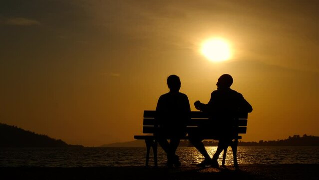 Traveling couple silhouette on evening bench. A view of couple silhouette relax on bench and admire the evening lake in summer.