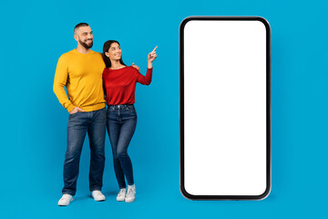 Smiling couple standing beside large phone and pointing at the blank screen